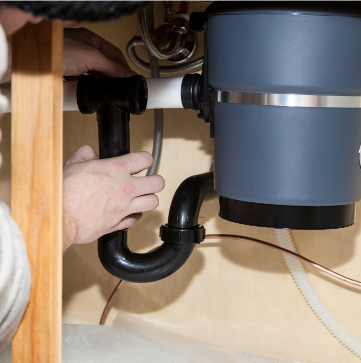 A close-up view of a technician installing a black drainage pipe assembly connected to a grey garbage disposal unit under a sink.